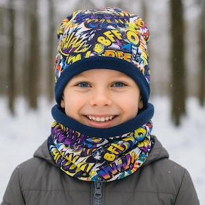 Op de afbeelding: Een kind draagt een kleurrijke wintermuts en nekwarmer set. De muts en nekwarmer hebben een graffiti-stijl ontwerp met het woord "BFF" zichtbaar. De muts en nekwarmer hebben een marineblauwe rand. Het kind draagt een grijze jas.