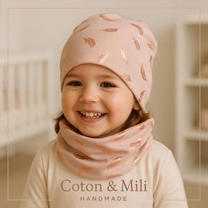 May include: A light pink beanie and neck warmer set with a feather pattern in a metallic rose gold color. The set is worn by a child. The text "Coton & Mili HANDMADE" is visible on the bottom of the image.