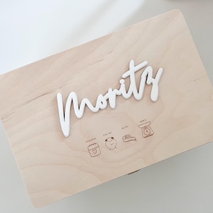Könnte beinhalten: Eine hellfarbene Holzkiste mit dem Namen "Moritz" in weißer Schrift. Die Kiste hat eingravierte Symbole und Text, die Geburtsdetails angeben. Die Kiste ist rechteckig und hat einen Klappdeckel.