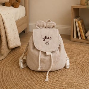Op de afbeelding: Een beige stoffen rugzak met twee ronde oren op de bovenste flap, een trekkoordsluiting en verstelbare schouderbanden. De flap is geborduurd met de naam Izhac in zwarte letters boven een kleine lijntekening van een teddybeer.
