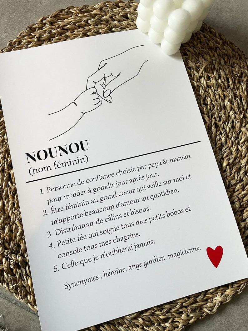 May include: A black and white line drawing of two hands holding each other. The text "NOUNOU (nom f&eacute;minin)" is at the top of the image. The text below the drawing defines the word "NOUNOU" as a person of trust chosen by parents to help a child grow. The text also describes the qualities of a "NOUNOU" as being a caring, loving, and supportive person. A red heart is in the bottom right corner of the image.