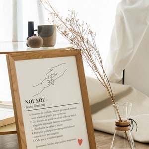 May include: A framed print with a black and white line drawing of two hands holding each other. The print has the word "NOUNOU" in large letters at the top and a definition of the word in French below the drawing. The definition includes the words "personne de confiance", "grand coeur", "c&acirc;lins", and "bisous".