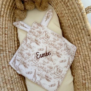 May include: A square baby security blanket with a white and brown toile print featuring giraffes, elephants, and airplanes. The blanket has two soft white bunny ears at the top and is embroidered with the name Esmée in brown script lettering.