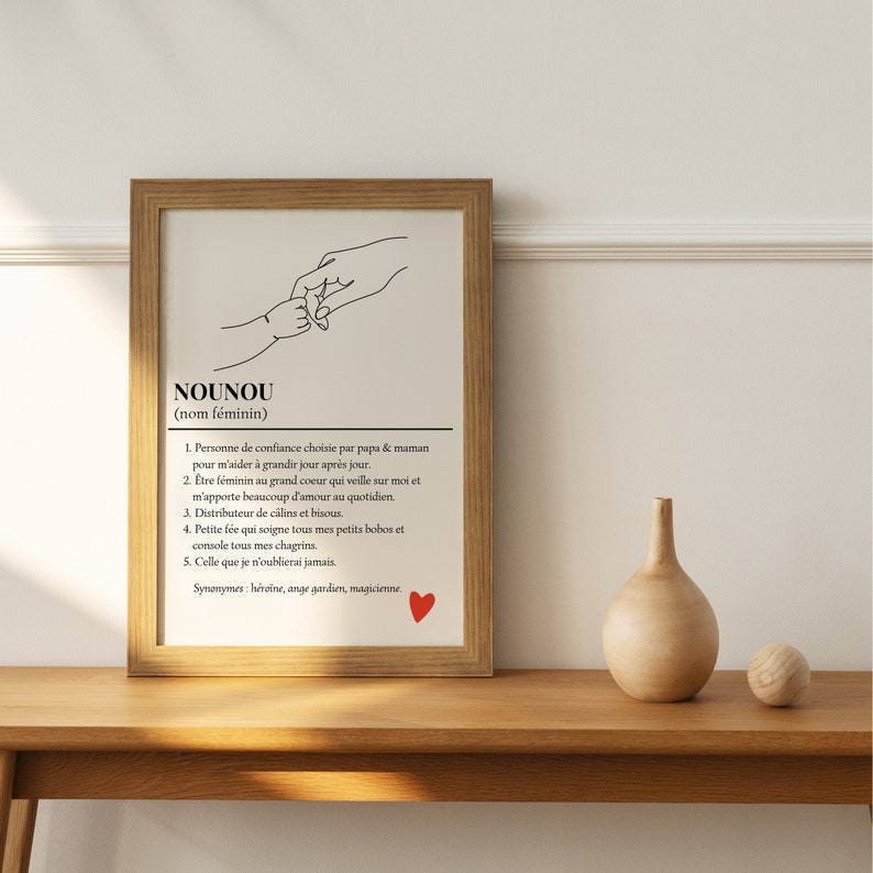 May include: Black and white line art illustration of two hands holding each other. The text below the image reads "NOUNOU (nom f&eacute;minin)" and defines the word as a person of trust chosen by parents to help a child grow. The text also describes the qualities of a nounou, such as being caring, supportive, and comforting. A red heart is in the bottom right corner of the image.
