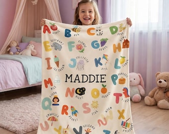 May include: A cream-colored fleece blanket featuring a colorful alphabet print with corresponding illustrations for each letter, such as A for Apple and B for Bee. The name MADDIE is printed in bold black capital letters in the center of the blanket.