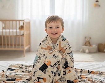 May include: A cream-colored blanket featuring a repeating pattern of construction vehicles, including excavators and dump trucks, in shades of blue, orange, and black. The name JAMES is printed in black text throughout the fabric design.