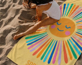 May include: A yellow beach towel featuring a large, smiling sun graphic with pink cheeks and radiating colorful rays in shades of blue, green, orange, and pink. The name Sophia is printed in bold orange capital letters at the bottom edge.