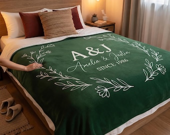 May include: A dark green fleece blanket featuring a white line-art floral wreath design. The center includes the white text A&J, Amelia & Justin, and SINCE 1986. The blanket is draped over a bed with white and dark red pillows and a textured throw pillow.