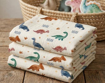 May include: A stack of three folded muslin blankets featuring a repeating pattern of watercolor dinosaurs in shades of blue, green, and brown, interspersed with small gold stars and the name Noah printed in a dark blue sans-serif font.