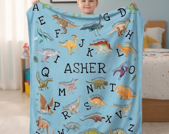May include: A light blue fleece blanket featuring the alphabet from A to Z, each letter paired with a dinosaur illustration and its name. The center of the blanket displays the name ASHER in large black capital letters. The design includes various colorful dinosaurs like a Triceratops, Pterodactyl, and Stegosaurus.