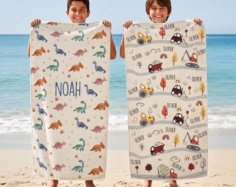 May include: Two rectangular beach towels with personalized prints. The left towel is cream-colored with a repeating pattern of colorful dinosaurs and the name NOAH in blue. The right towel features construction vehicles and the name OLIVER.