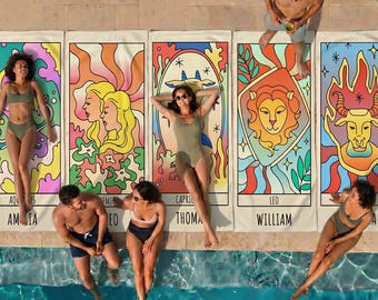 May include: Five rectangular beach towels featuring colorful zodiac-themed illustrations with names printed at the bottom, laid out on a stone pool deck. The designs include Gemini, Capricorn, Leo, and Taurus motifs in vibrant, psychedelic patterns.