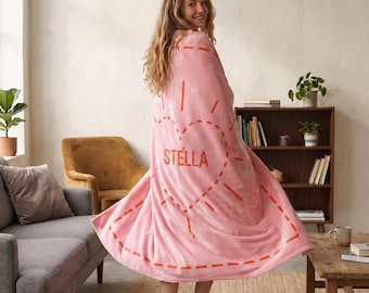May include: A soft pink fleece blanket featuring a large orange dashed heart outline and the name STELLA printed in orange capital letters in the center. The blanket is draped to show the full design, which includes orange dashed lines radiating outward.