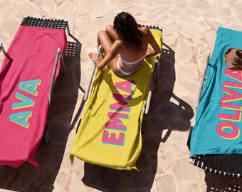 May include: Three rectangular beach towels draped over lounge chairs. The left towel is pink with the name AVA in yellow and blue. The center towel is yellow with the name EMMA in pink and blue. The right towel is blue with the name OLIVIA in pink and yellow.