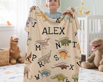 May include: A cream-colored fleece blanket featuring a dinosaur alphabet print with the name ALEX in large black letters. The design includes various colorful dinosaur illustrations paired with corresponding letters of the alphabet in a bold black font.