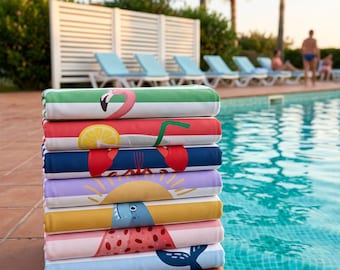 May include: A stack of seven folded rectangular beach towels, each featuring a distinct graphic print including a pink flamingo, a lemon slice, a red lobster, a yellow sun, a blue shark, a watermelon slice, and a dark blue whale tail.