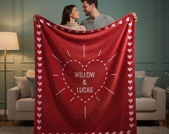 May include: A deep red rectangular blanket featuring a central dashed-line heart graphic with the text WILLOW & LUCAS inside. The border is decorated with a repeating pattern of small light pink hearts along all four edges of the fabric.