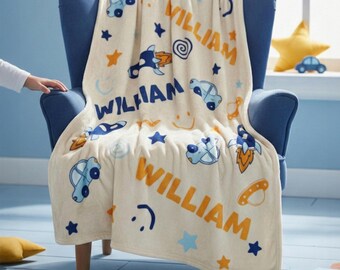 May include: A cream-colored fleece blanket featuring a repeating pattern of blue cars, orange rockets, yellow stars, and smiley faces. The name WILLIAM is printed in both blue and orange text throughout the design, creating a playful, colorful aesthetic.