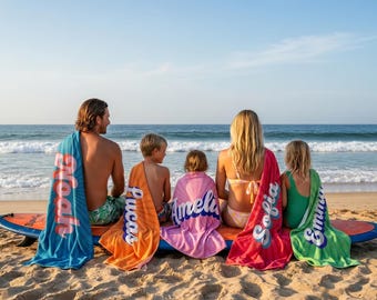 May include: Five rectangular beach towels in blue, orange, pink, red, and green, each featuring a large, white, cursive name printed vertically. The towels are draped over a long surfboard resting on the sand near the ocean shoreline.