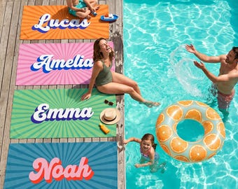 May include: Four rectangular beach towels with sunburst patterns in orange, pink, green, and blue. Each towel features a large, white, retro-style name printed in the center: Lucas, Amelia, Emma, and Noah. The towels are laid out on a wooden deck.