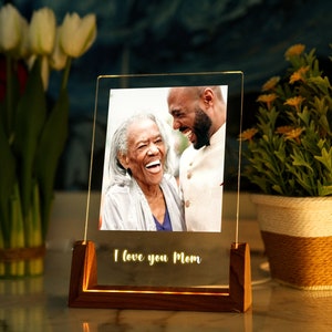 Puede incluir: Un marco de fotos acr&iacute;lico iluminado con una base de madera. El marco muestra una fotograf&iacute;a de dos personas riendo. Las palabras "I love you Mom" est&aacute;n iluminadas en la parte inferior. Tulipanes y una planta en maceta est&aacute;n en el fondo.