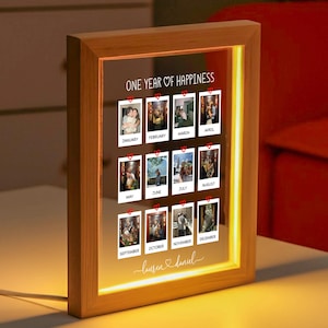 May include: A wooden framed photo display with a warm, glowing light. The display features twelve small photos, each representing a month, with the text "ONE YEAR OF HAPPINESS" at the top. The names "Lauren & Daniel" are written at the bottom.