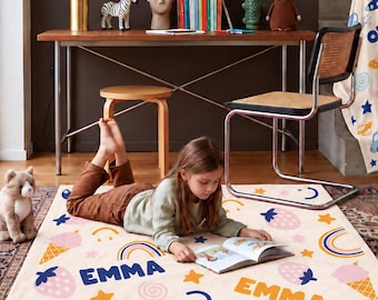 May include: A cream-colored rectangular blanket featuring the name EMMA in blue and orange text, decorated with pink strawberries, orange and blue rainbows, ice cream cones, yellow stars, and blue smiley faces, spread out on a patterned rug.