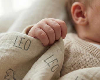 May include: A beige muslin swaddle blanket featuring the name LEO printed in a dark gray sans-serif font, accompanied by a small, simple line-art illustration of a dinosaur. The fabric has a soft, textured, and lightweight appearance.