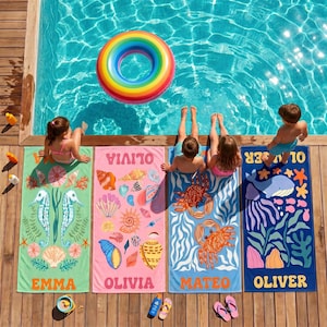 May include: Four colorful beach towels with personalized names and sea-themed designs are laid out on a wooden deck next to a pool. A rainbow-colored inflatable ring floats in the pool. The towels feature seahorses, shells, crabs, and a whale.