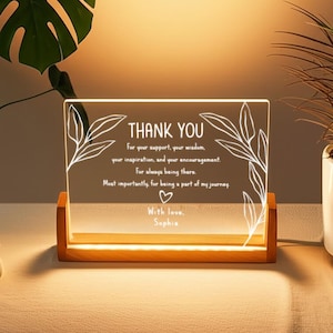 Può includere: Una placca rettangolare in acrilico illuminata con la scritta "THANK YOU" e un messaggio personalizzato. La placca è sostenuta da una base in legno. Disegni di foglie bianche incorniciano il testo, che include un simbolo a cuore e il nome "Sophia".