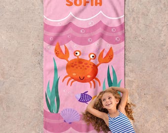 May include: A rectangular beach towel featuring a pink background with a repeating wave pattern, white bubbles, green seaweed, a purple seashell, a small purple fish, and a large orange crab. The name SOFIA is printed in orange at the top.