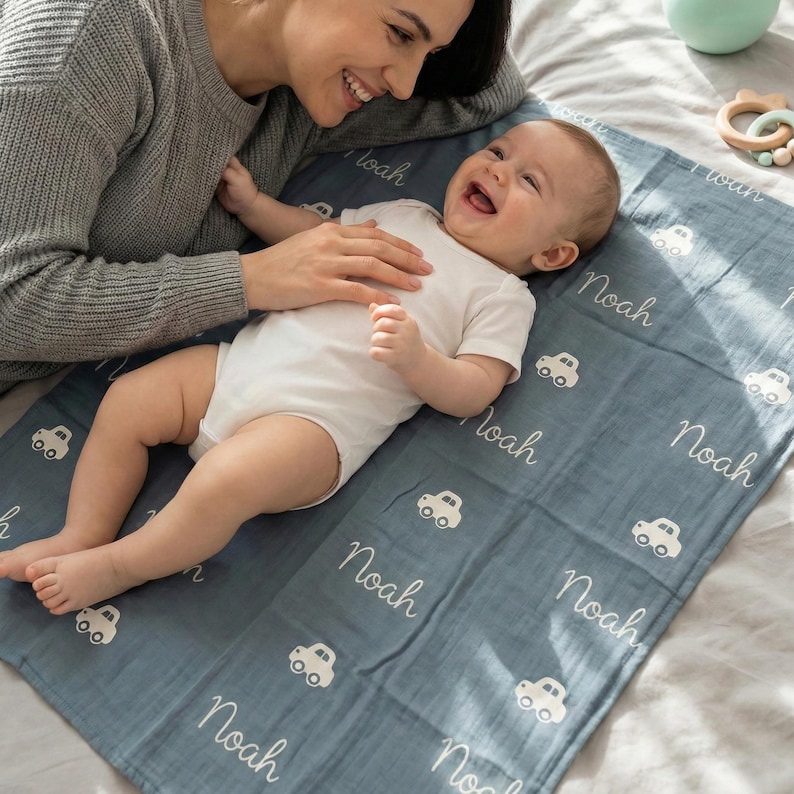 May include: A baby lies on a blue blanket with the name "Noah" and white car illustrations. The baby is wearing a white onesie. A smiling person is leaning over the baby, touching the blanket. A green bottle and wooden toy are in the background.