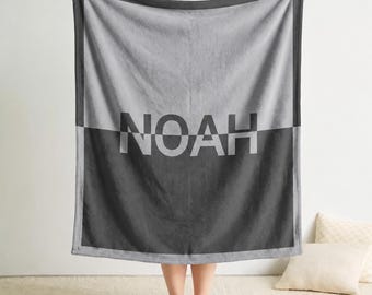 May include: A rectangular fleece blanket featuring a two-tone design with a light gray top half and a dark charcoal gray bottom half. The name NOAH is printed in the center in a split-color font, with the top half in dark gray and the bottom half in light gray.