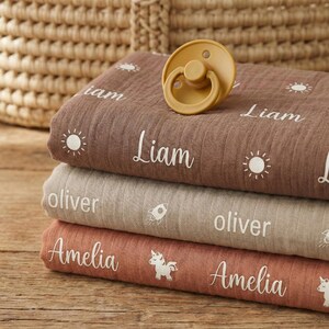May include: A stack of three personalized baby blankets in shades of brown, tan, and coral. Each blanket features a name in white script, along with a sun or rocket graphic. A mustard-colored pacifier rests on top.