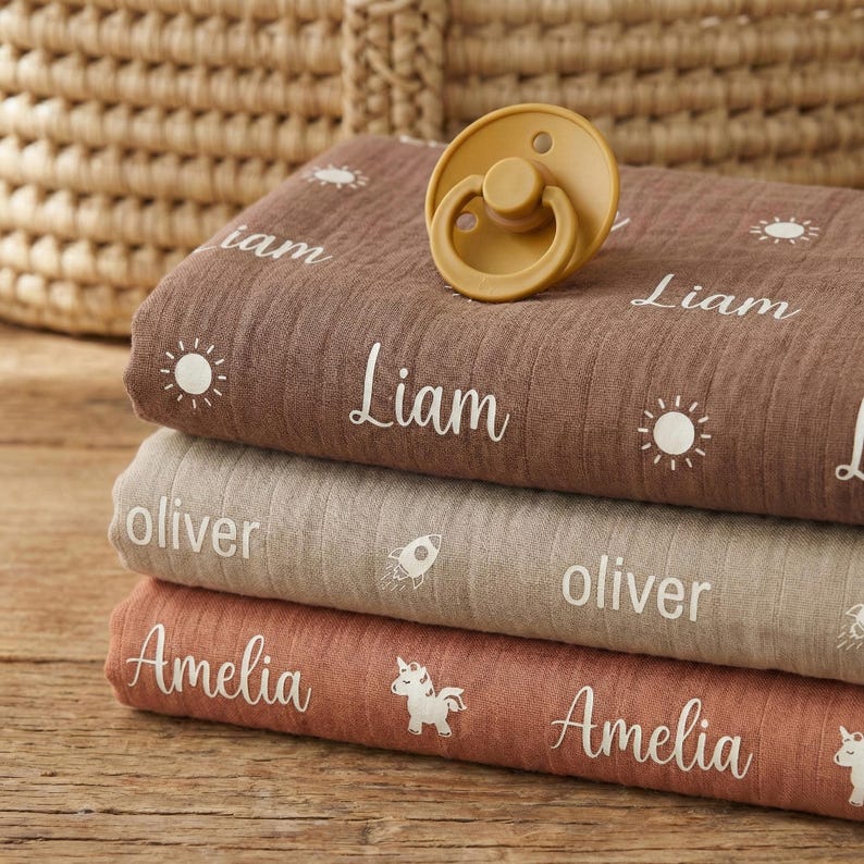 May include: A stack of three personalized baby blankets in shades of brown, tan, and coral. Each blanket features a name in white script, along with a sun or rocket graphic. A mustard-colored pacifier rests on top.