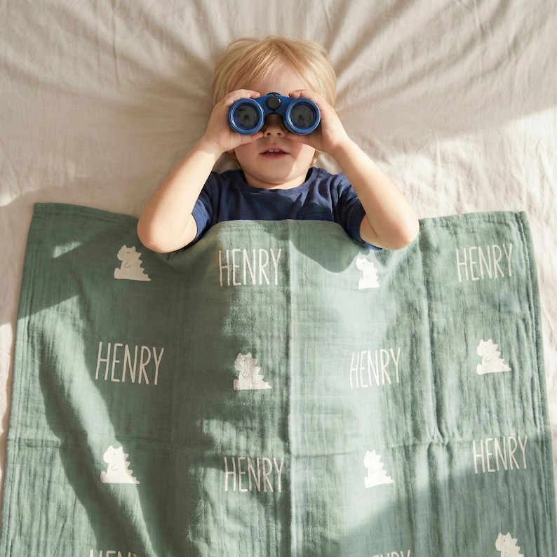 May include: A young child lies on a bed, looking through blue binoculars. A light green blanket with the name "HENRY" and dinosaur illustrations is partially covering the child. The blanket is made of a soft, textured fabric.