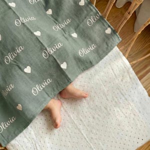 May include: A sage green baby blanket with the name "Olivia" and white hearts printed across the fabric. The blanket covers a baby's feet, resting on a white dotted sheet. The blanket is made of soft material.