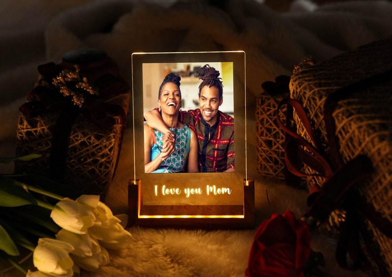 Puede incluir: Marco de fotos iluminado de madera con una foto de una pareja sonriendo. La base muestra las palabras "I love you Mom". El fondo incluye regalos y flores, creando un ambiente c&aacute;lido.