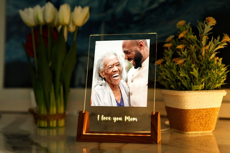 Puede incluir: Un marco de fotos acr&iacute;lico iluminado con una base de madera. El marco muestra una fotograf&iacute;a de dos personas riendo. Las palabras "I love you Mom" est&aacute;n iluminadas en la parte inferior. Tulipanes y una planta en maceta est&aacute;n en el fondo.