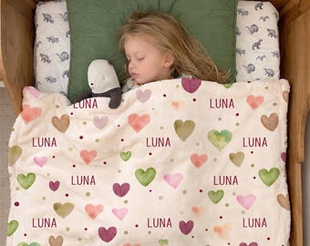 May include: A soft cream-colored fleece blanket featuring a repeating pattern of watercolor hearts in shades of pink, green, and brown, interspersed with small dark red dots and the name LUNA printed in a dark red sans-serif font throughout.