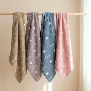 May include: Four personalized baby blankets in shades of tan, purple, blue, and pink, hanging on a wooden rod. Each blanket features a name and a pattern of white stars, hearts, or cars. The names include Henry, Charlotte, Noah, and Sophia.