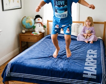 May include: A dark blue plush blanket featuring a white border and the name HARPER printed in white block letters. The blanket is spread across a wooden bed frame, providing a soft surface for a child jumping in winter-themed pajamas.