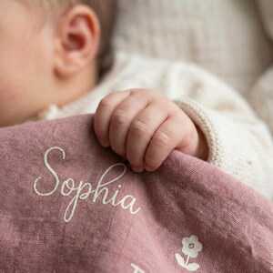 May include: A dusty rose-colored baby blanket with the name "Sophia" embroidered in a delicate script. A small floral design is visible near the bottom. The blanket is soft and appears to be made of a lightweight fabric.