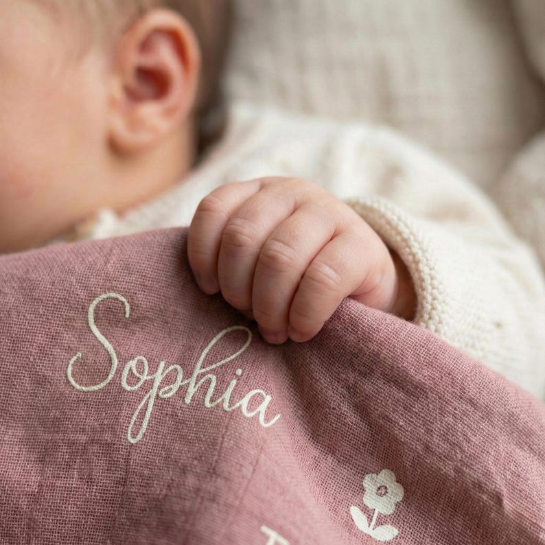 May include: A dusty rose-colored baby blanket with the name "Sophia" embroidered in a delicate script. A small floral design is visible near the bottom. The blanket is soft and appears to be made of a lightweight fabric.