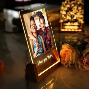 Puede incluir: Un marco de fotos iluminado con una foto de una pareja sonriendo. El marco es de acr&iacute;lico transparente y se apoya en una base de madera. Las palabras "I love you Mom" est&aacute;n iluminadas. Rosas y una peque&ntilde;a calabaza en el fondo.