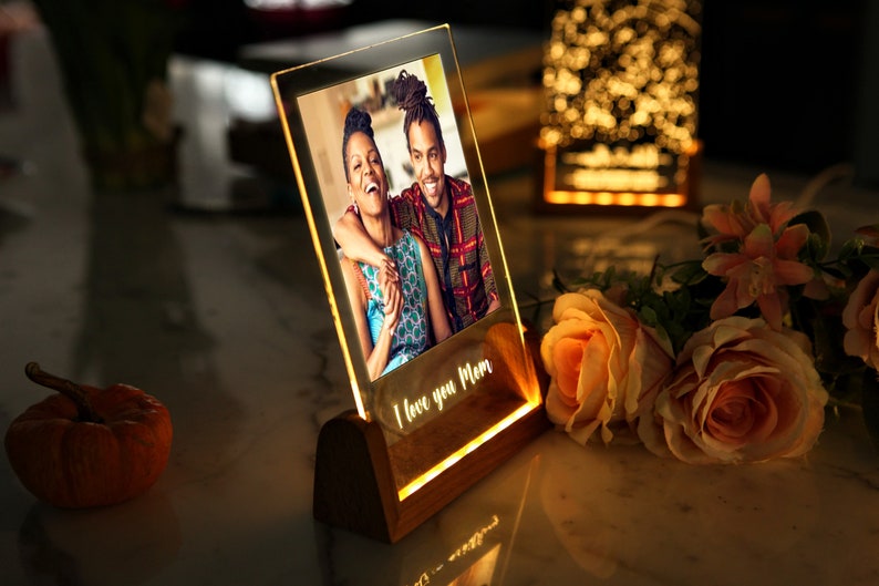 Puede incluir: Un marco de fotos iluminado con una foto de una pareja sonriendo. El marco es de acr&iacute;lico transparente y se apoya en una base de madera. Las palabras "I love you Mom" est&aacute;n iluminadas. Rosas y una peque&ntilde;a calabaza en el fondo.