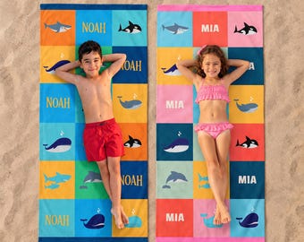 May include: Two rectangular beach towels featuring a grid pattern of colorful squares. One towel displays the name NOAH with illustrations of sharks and whales. The other towel displays the name MIA with similar marine life graphics.