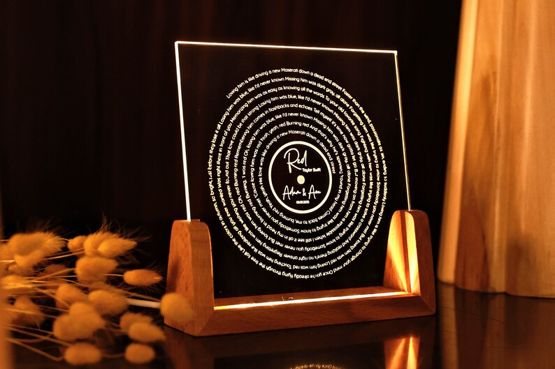 May include: A wooden stand with a clear acrylic panel displaying a circular design of song lyrics. The lyrics are in a white font and the centre of the design features a record with the text "Red River Blue" and "Adam & Ava" in a gold font.