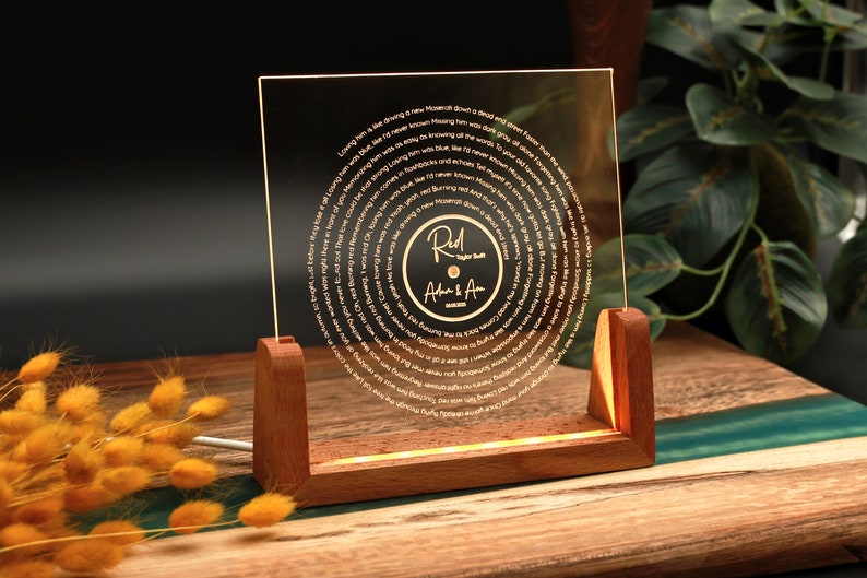 May include: A clear acrylic record with a gold inscription of song lyrics in a spiral pattern. The record is on a wooden stand with a light shining through the acrylic. The record has a gold label with the text "Red" and "Adam & Ava" in cursive.