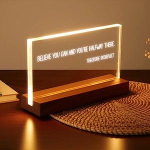 Puede incluir: Un letrero acrílico iluminado con la cita "BELIEVE YOU CAN AND YOU'RE HALFWAY THERE" de Theodore Roosevelt. El letrero está iluminado con un brillo cálido y se asienta sobre una base de madera. El fondo incluye un mantel individual tejido y un libro.